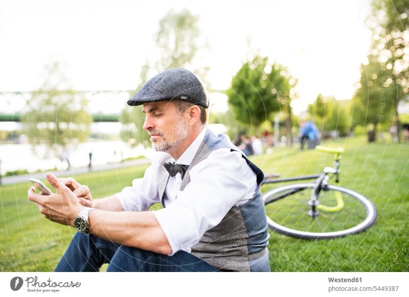 Confident mature businessman with bicycle and smartphone in the city park sitting on grass mobile phone mobiles mobile phones Cellphone cell phone cell phones