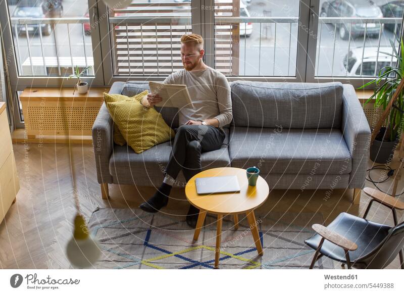 Man sitting on couch at home reading newspaper newspapers settee sofa sofas couches settees man men males Seated Adults grown-ups grownups adult people persons