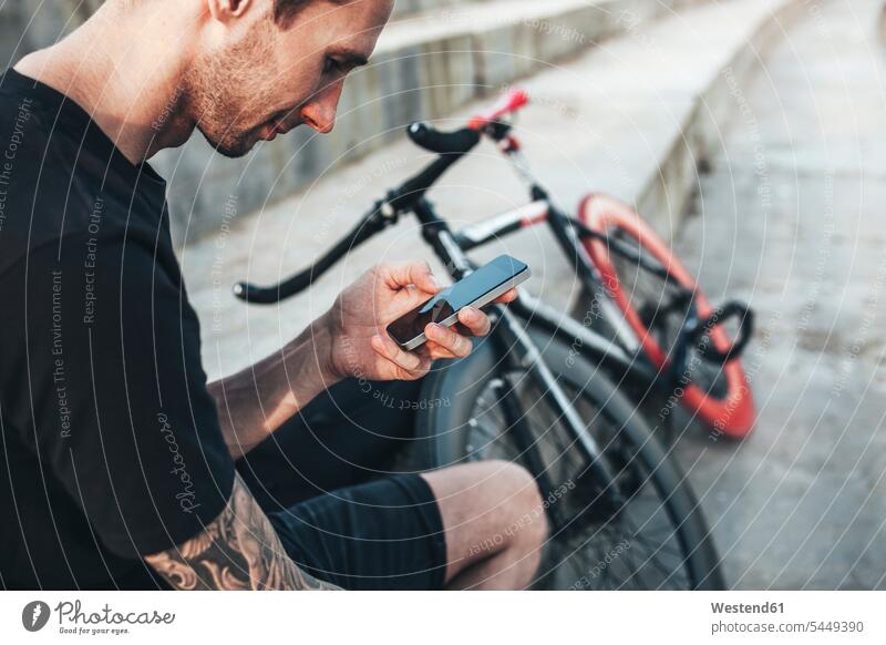 Young man sitting next to fixie bike using cell phone Seated men males bicycle bikes bicycles mobile phone mobiles mobile phones Cellphone cell phones Adults