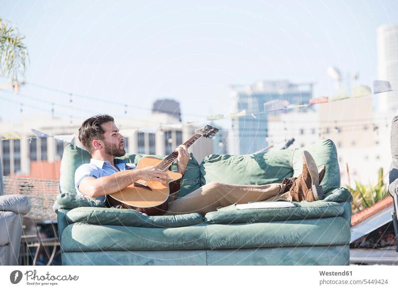 Young man on rooftop lying on sofa and playing guitar couch settee sofas couches settees musician musicians laying down lie lying down roof terrace deck guitars