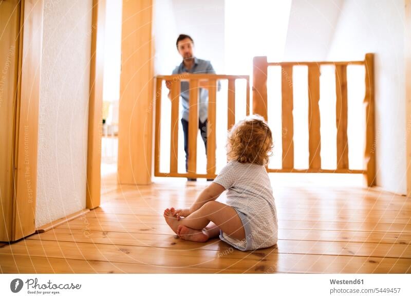 Baby boy sitting on wooden floor looking at father behind barrier at stairs baby infants nurselings babies stairway barriers home at home pa fathers daddy dads
