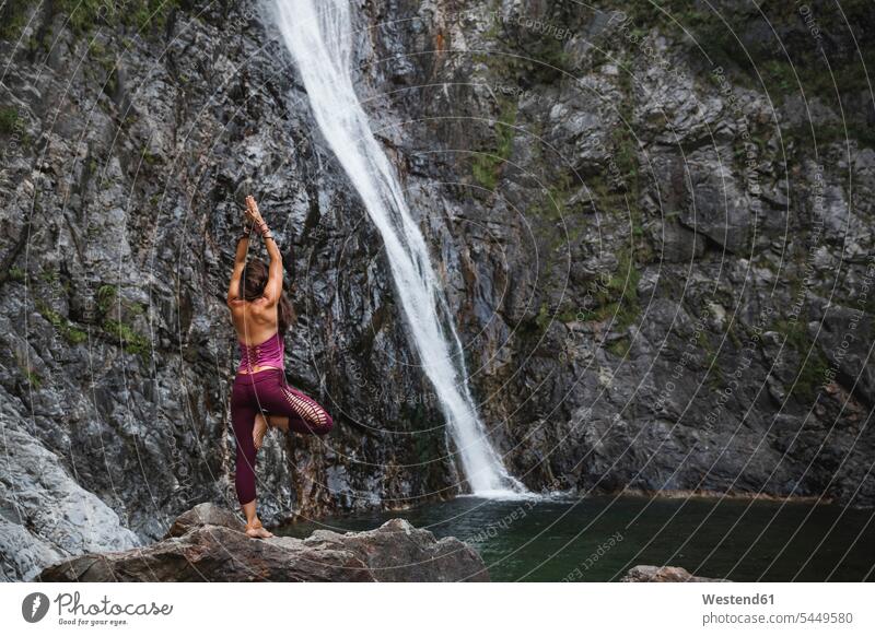 Italy, Lecco, woman doing Tree Yoga Pose on a rock near a waterfall waterfalls females women yoga standing waters body of water Adults grown-ups grownups adult