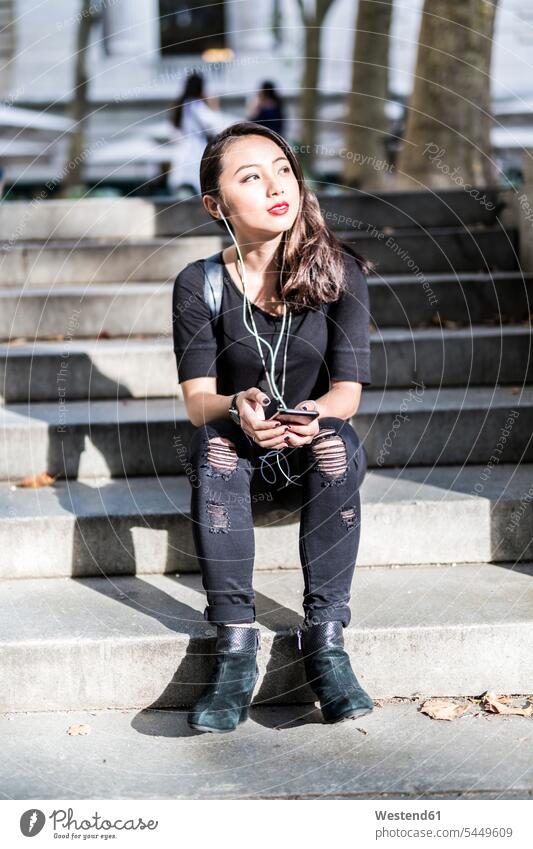 Portrait of young woman dressed in black sitting on steps listening music with earphones and cell phone females women Adults grown-ups grownups adult people