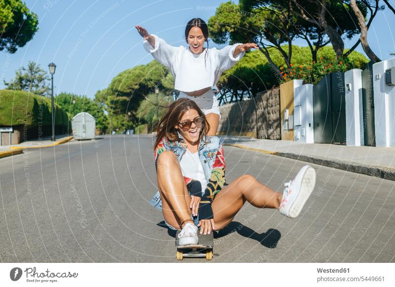 Two laughing friends together on skateboard female friends Skate Board skateboards road streets roads Laughter mate friendship positive Emotion Feeling Feelings