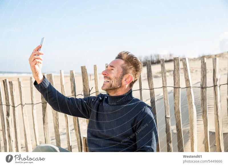 Man taking a selfie at fence on the beach sitting Seated beaches mobile phone mobiles mobile phones Cellphone cell phone cell phones Selfie Selfies man men