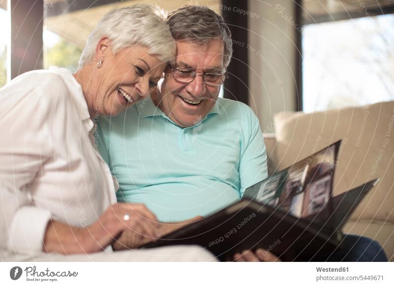 Happy senior couple sitting on couch looking at photo album settee sofa sofas couches settees Seated eyeing happiness happy twosomes partnership couples home