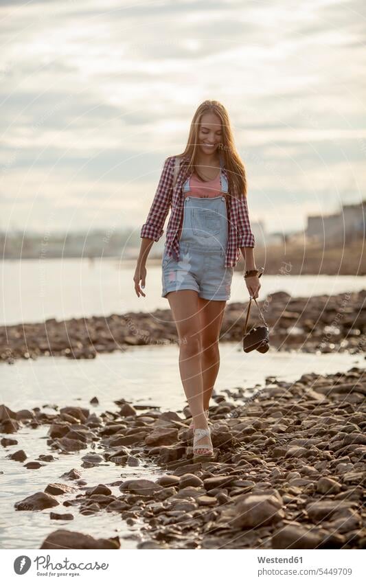 Young woman holding a camera walking on stony beach River Rivers going cameras smiling smile females women water waters body of water Adults grown-ups grownups