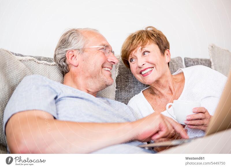Happy senior couple at home sitting on couch using laptop settee sofa sofas couches settees Laptop Computers laptops notebook smiling smile relaxed relaxation