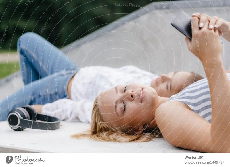 Two young women with cell phone and headphones lying on ramp in a skatepark female friends headset Skateboard Park skate park laying down lie lying down parks