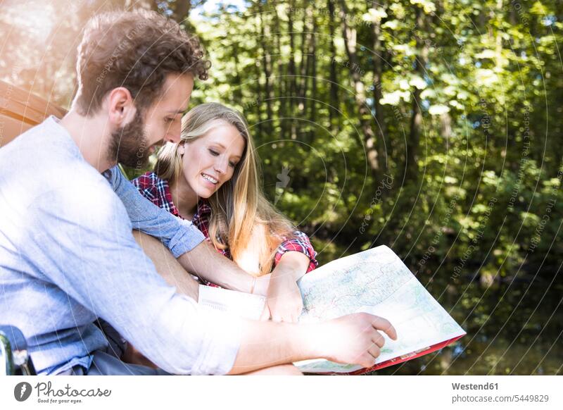 Smiling young couple reading map at a brook brooks rivulet twosomes partnership couples forest woods forests smiling smile looking eyeing maps water waters