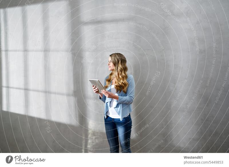 Blond woman with tablet in front of grey wall looking at distance females women Adults grown-ups grownups adult people persons human being humans human beings