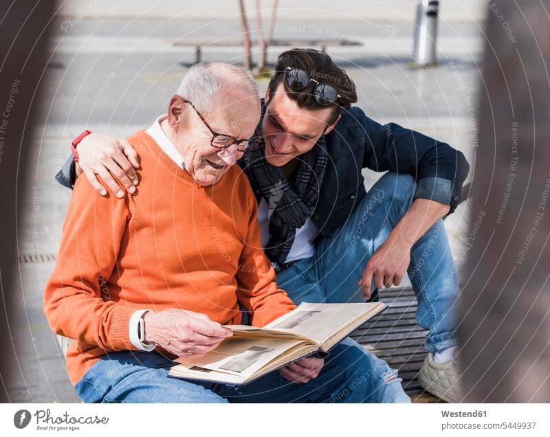 Senior man and adult grandson on a bench looking at photo album grandsons sitting Seated benches eyeing smiling smile photograph album photo albums grandfather