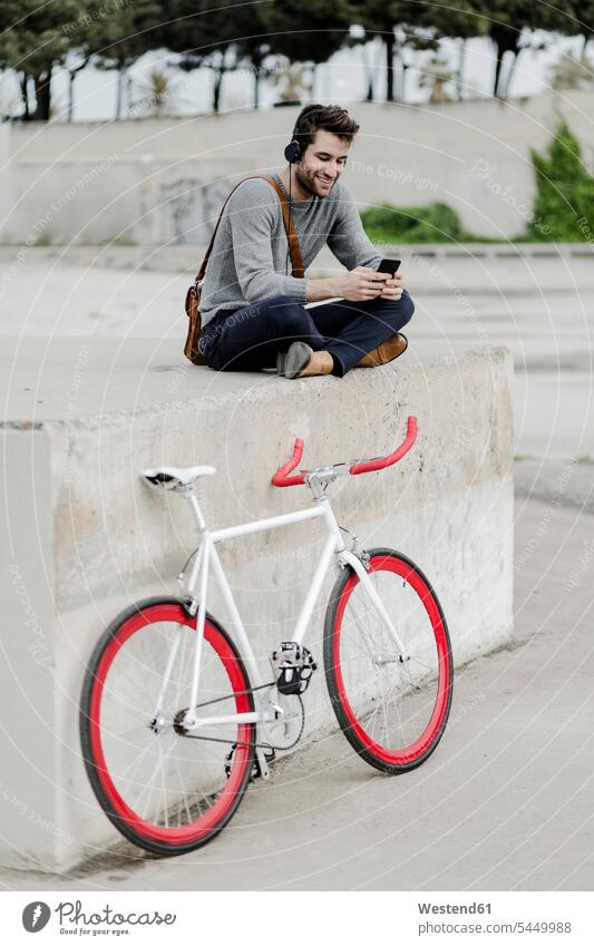 Young man with racing cycle sitting on a wall listening music with headphones men males headset Smartphone iPhone Smartphones Adults grown-ups grownups adult