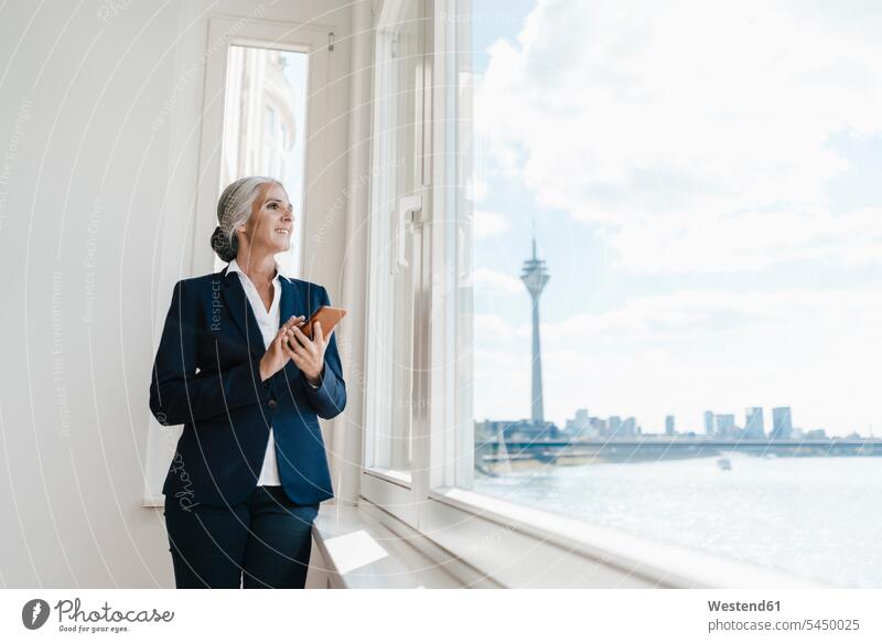 Businesswoman looking out of window in waterfront office businesswoman businesswomen business woman business women smiling smile windows standing mobile phone