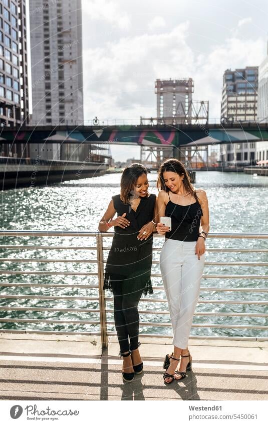 Two women on a bridge sharing a cell phone in the city woman females Female Colleague smiling smile mobile phone mobiles mobile phones Cellphone cell phones