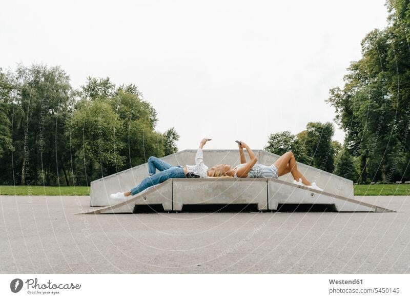 Two young women with cell phones lying on ramp in a skatepark female friends woman females Ramp Skateboard Park skate park laying down lie lying down