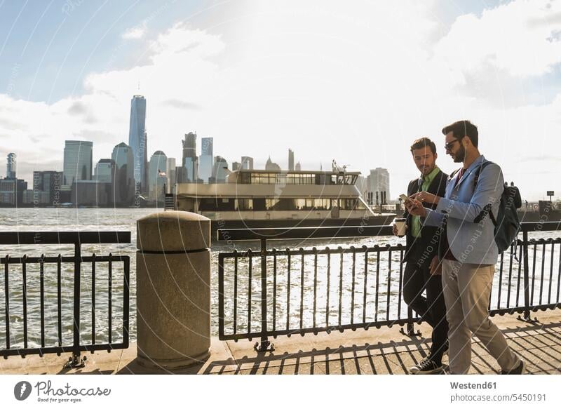 USA, two businessmen walking at New Jersey waterfront with view to Manhattan New York State Businessman Business man Businessmen Business men going mobile phone