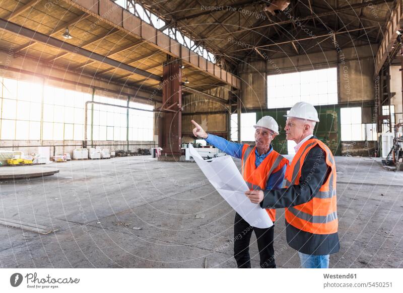 Two men with plan wearing safety vests talking in old industrial hall construction plan building plan architectural drawing shop floor factory hall
