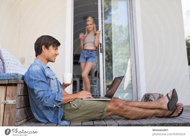 Man sitting on terrace using laptop with woman in background Laptop Computers laptops notebook men males smiling smile terraces Seated computer computers Adults