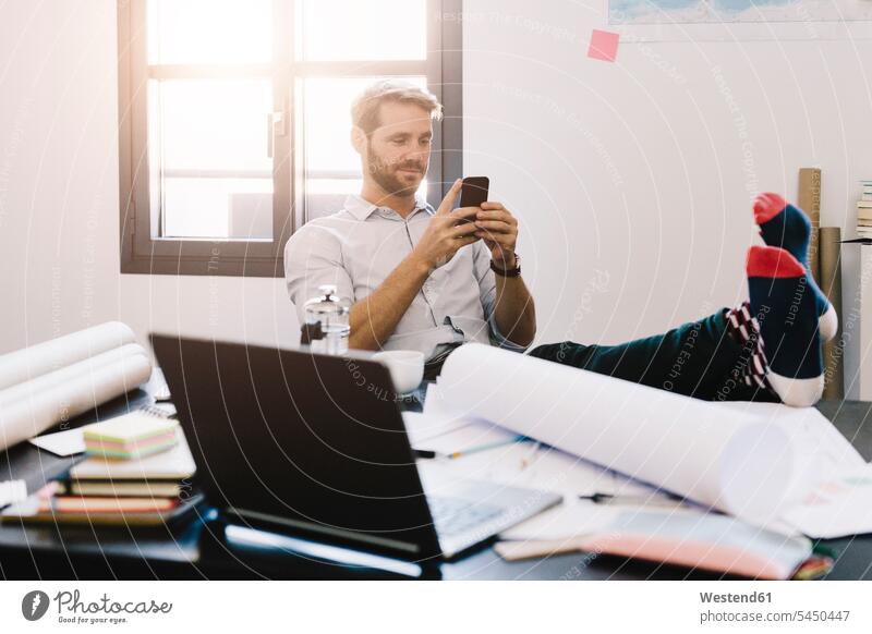 Portrait of smiling architect sitting with feet up at desk using cell phone office offices office room office rooms architects workplace work place