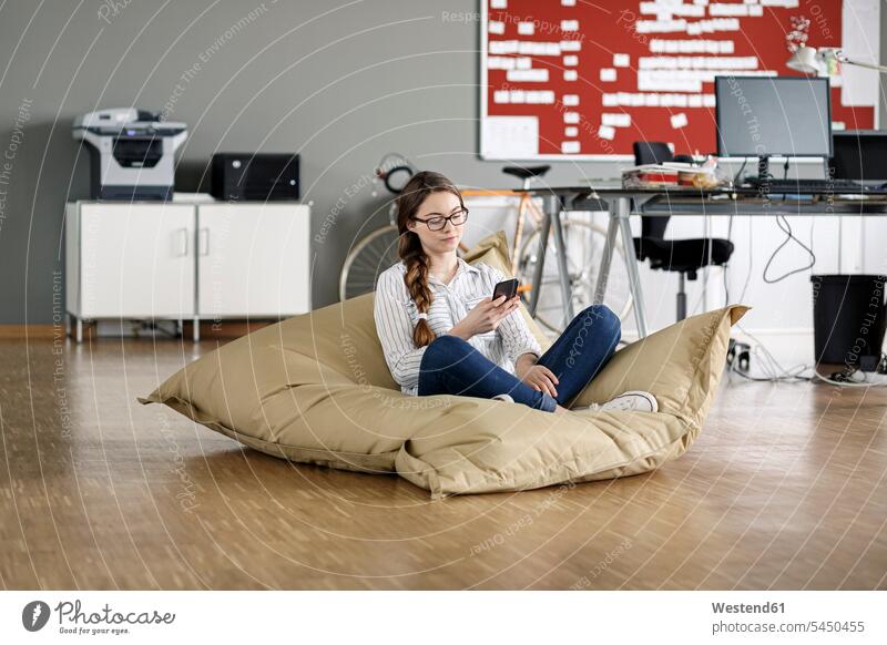 Young woman with cell phone sitting in bean bag in office offices office room office rooms Seated mobile phone mobiles mobile phones Cellphone cell phones