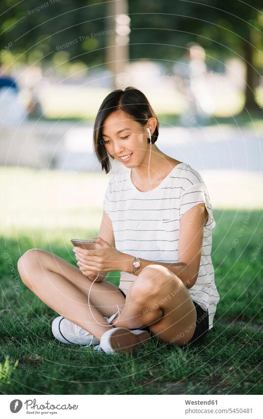 Smiling young woman sitting on a meadow listening music with earphones females women Adults grown-ups grownups adult people persons human being humans