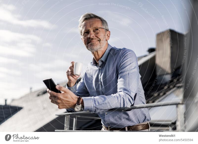 Grey-haired businessman with smartphone standing on balkony drinking coffee Coffee smiling smile Businessman Business man Businessmen Business men mobile phone