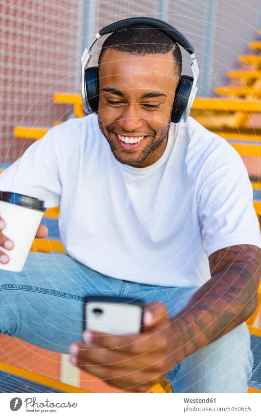 Laughing young man with headphones and coffee to go sitting on stairs looking at smartphone men males Smartphone iPhone Smartphones headset Adults grown-ups