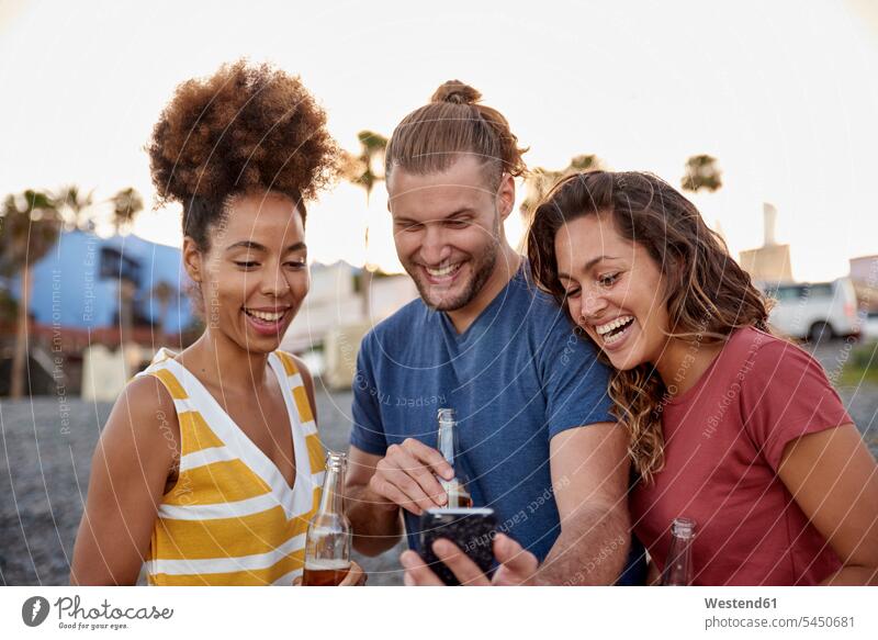 Three laughing friends with beer bottles looking at smartphone on the beach beaches Selfie Selfies friendship Smartphone iPhone Smartphones Beer Bottle