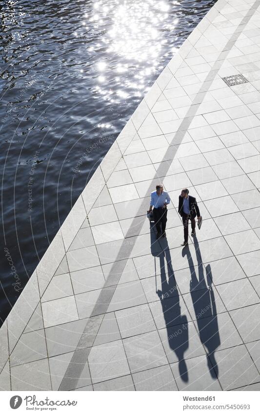 Two businessmen walking and talking at the riverbank speaking going Businessman Business man Businessmen Business men colleagues business people businesspeople