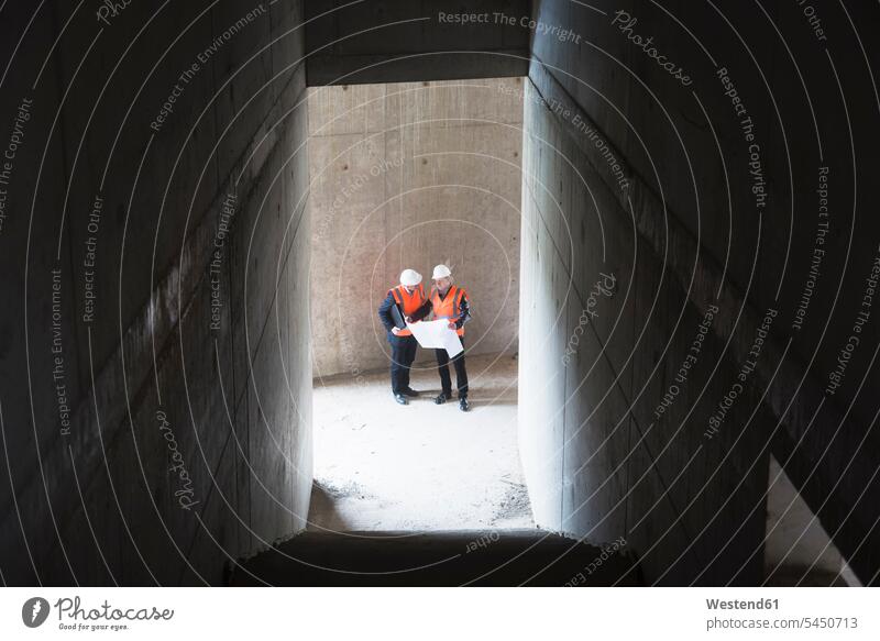 Two men with plan wearing safety vests talking in building under construction speaking working At Work man males architect architects construction site