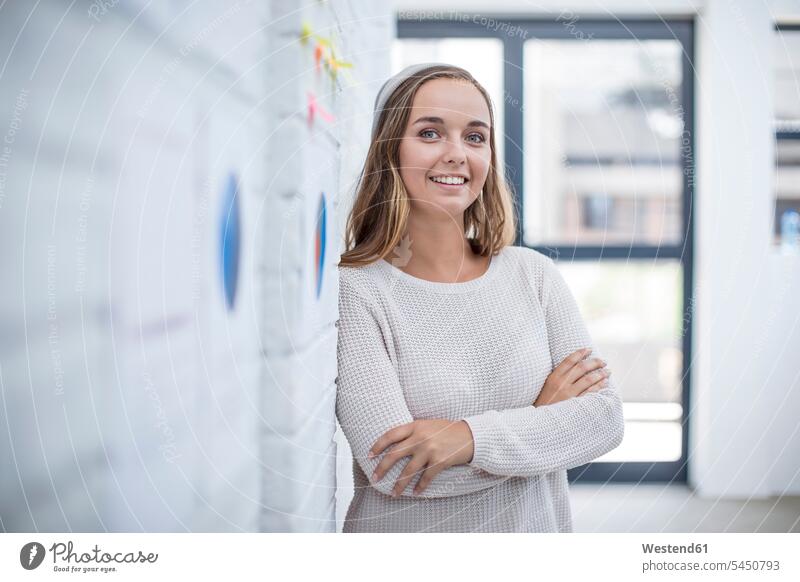 Young woman leaning against wall in office with warmd crossed walls office worker smiling smile Office Offices confidence confident white collar worker clerk