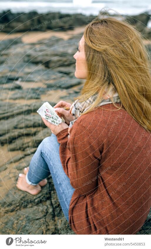 Woman with notebook sitting on rocks on the beach in winter woman females women notebooks Seated beaches Adults grown-ups grownups adult people persons