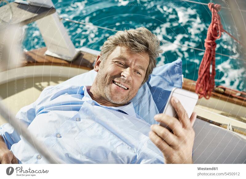Portrait of smiling mature man lying on deck of his sailing boat looking at cell phone men males portrait portraits Smartphone iPhone Smartphones Adults