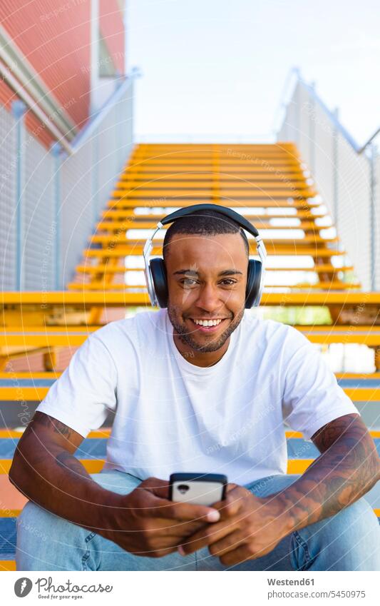 Portrait of laughing young man with headphones and smartphone sitting on stairs headset portrait portraits Smartphone iPhone Smartphones men males mobile phone