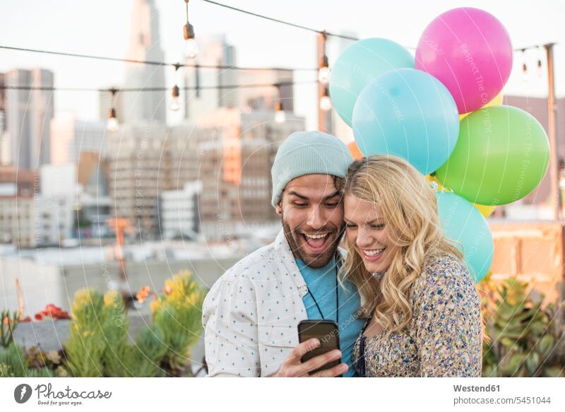 Young couple with balloons standing on rooftop terrace, using smart phone watching looking looking at Smartphone iPhone Smartphones twosomes partnership couples