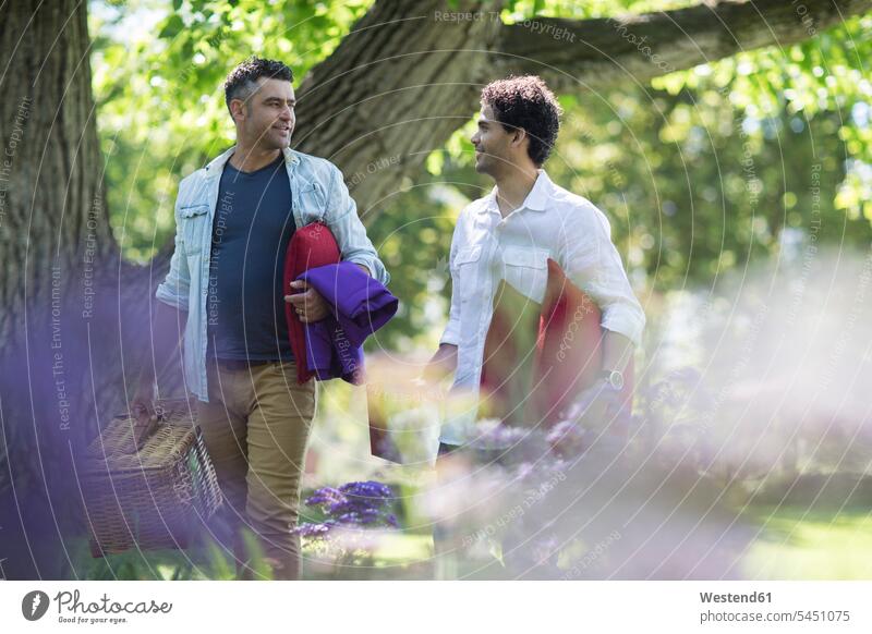 Two friends walking in park with picnic basket and cushions going man men males smiling smile Adults grown-ups grownups adult people persons human being humans