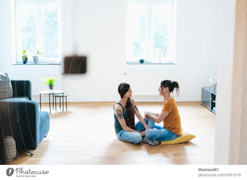 Two women sitting on wooden floor at home talking Seated tattoo tattoos woman females speaking tattooed body art Body Adornment Skin Art style stylish Adults