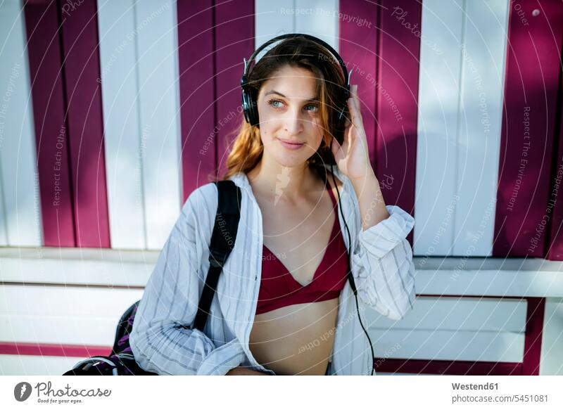 Young woman listening to music with headphones in front of wall headset females women hearing smiling smile Adults grown-ups grownups adult people persons