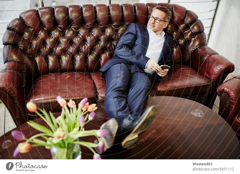 Mature businessman on couch holding cell phone mobile phone mobiles mobile phones Cellphone cell phones settee sofa sofas couches settees Businessman