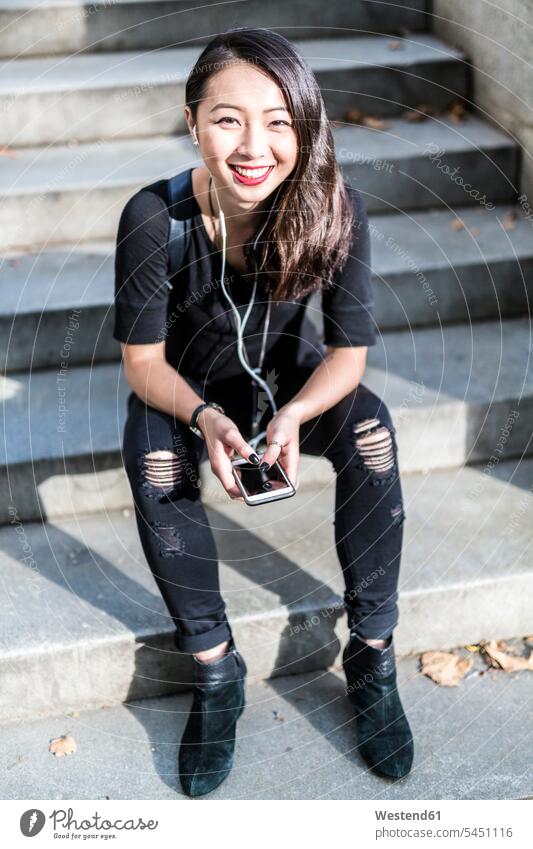 Portrait of young woman dressed in black sitting on steps listening music with earphones and cell phone smiling smile females women Adults grown-ups grownups