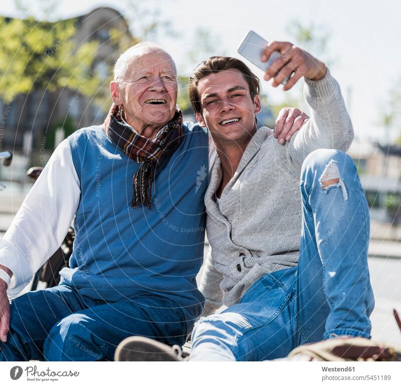 Senior man and adult grandson on a bench taking a selfie Fun having fun funny happiness happy sitting Seated smiling smile grandsons Selfie Selfies benches