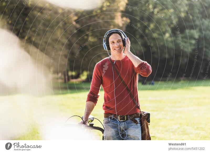 Laughing man with racing cycle listening music with headphones in a park men males headset parks Adults grown-ups grownups adult people persons human being