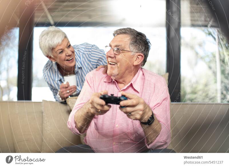 Happy senior man with wife playing video game on couch at home settee sofa sofas couches settees happiness happy video games couple twosomes partnership couples