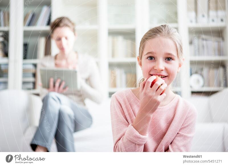 Girl eating apple at home with mother in background girl females girls smiling smile family families Apple Apples child children kid kids people persons