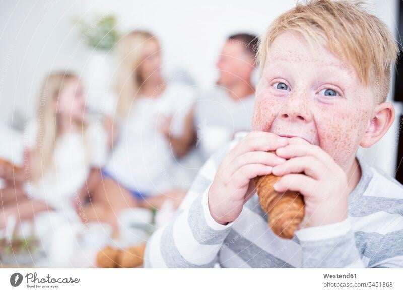 Portrait of boy biting into croissant with family in background Croissant Croissants Cornetto Cornettos Breakfast eating boys males Food foods food and drink