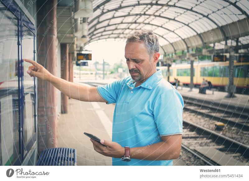 Man with smartphone standing on platform in front of timetable train station man men males Adults grown-ups grownups adult people persons human being humans