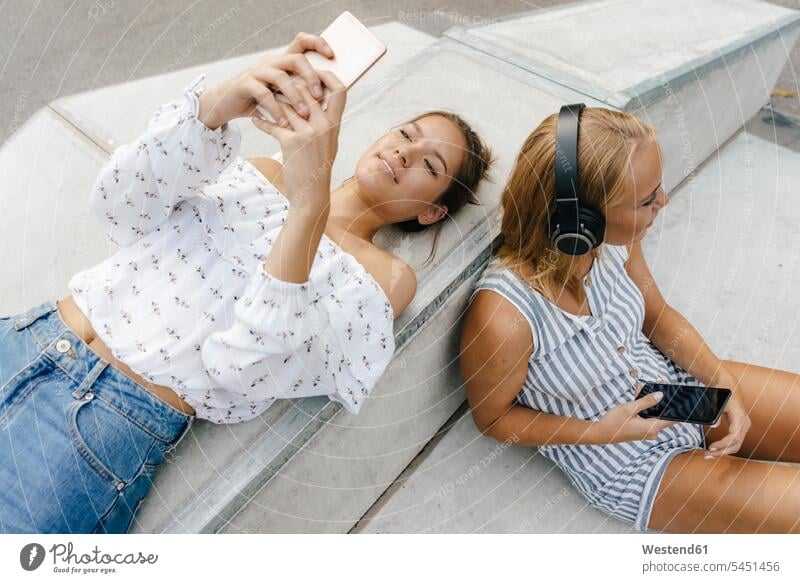Two young women with cell phones and headphones in a skatepark female friends headset mobile phone mobiles mobile phones Cellphone woman females Skateboard Park