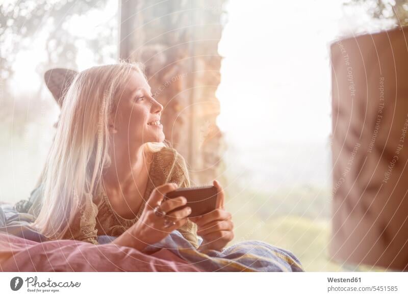 Portrait of smiling young woman lying on bed with cell phone looking through window females women Adults grown-ups grownups adult people persons human being