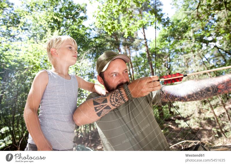 Son watching father shooting with bow and arrow in the forest son sons manchild manchildren woods forests arrows pa fathers daddy dads papa aiming boy boys